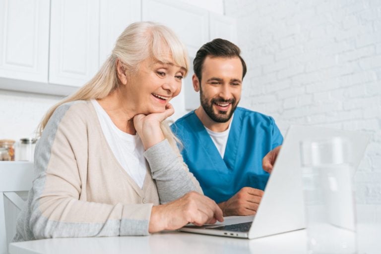 smiling young caregiver and happy senior woman using laptop together