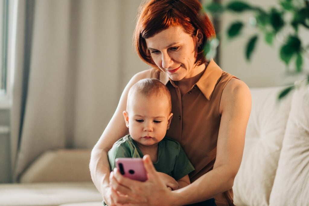 Childcare nanny holding infant while using mobile app, concept of updating communication with parent