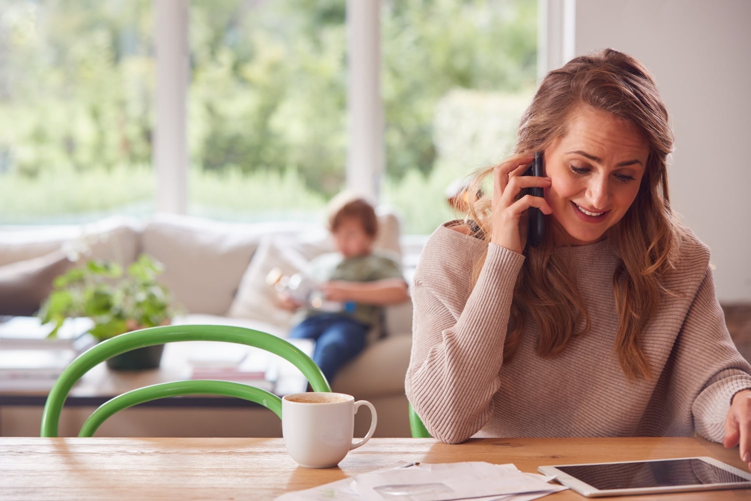mother sits at a wooden table, talking on her phone. young child sits on a sofa in the background