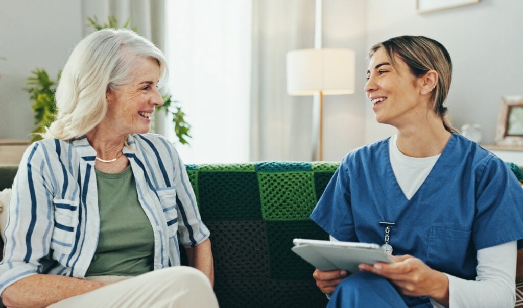 smiling senior woman sits while talking with a caregiver holding a tablet, related to home care, senior care, healthcare, nursing